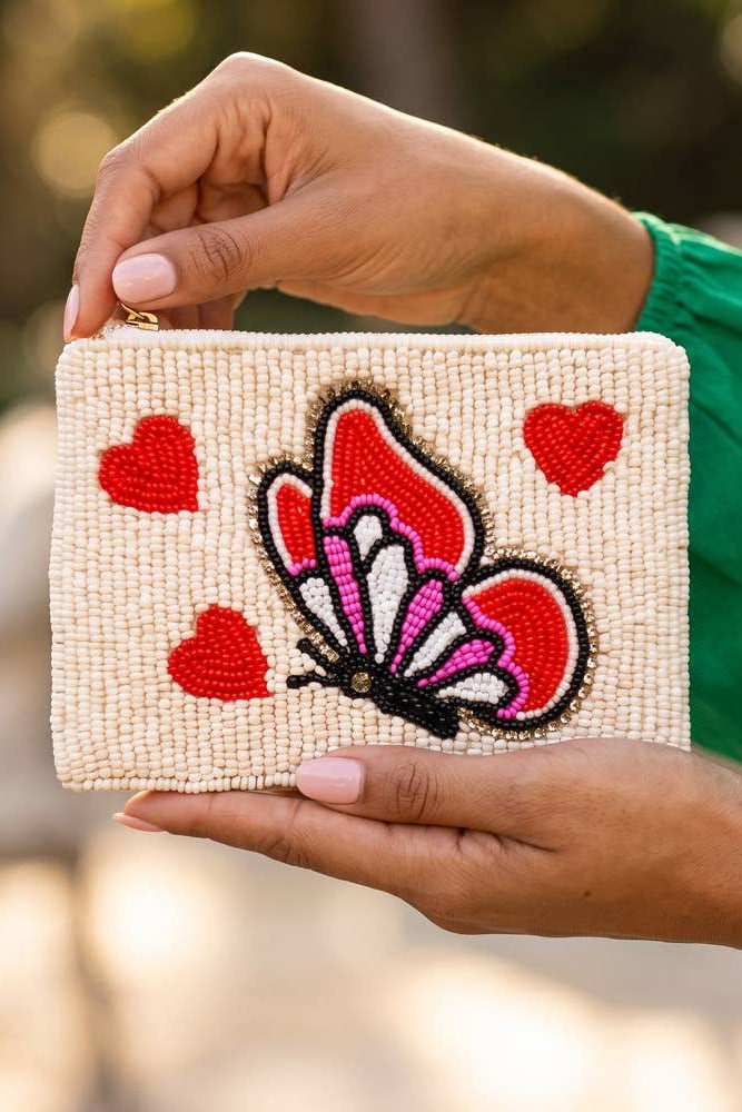 A person holds a small rectangular beaded clutch bag with a cream base, red hearts, and a red, pink, and white butterfly design. The person is wearing a bright green top, and the background is a softly blurred outdoor setting.