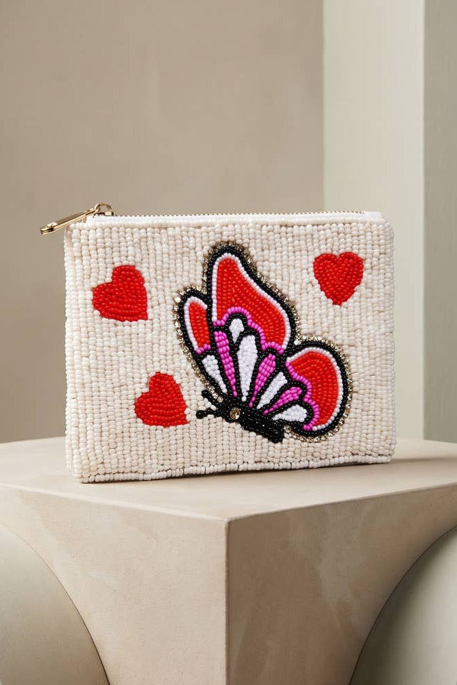 A small woven pouch with a gold zipper, featuring a central embroidered butterfly and three red hearts on a cream textured background, displayed on a beige sculptural pedestal.