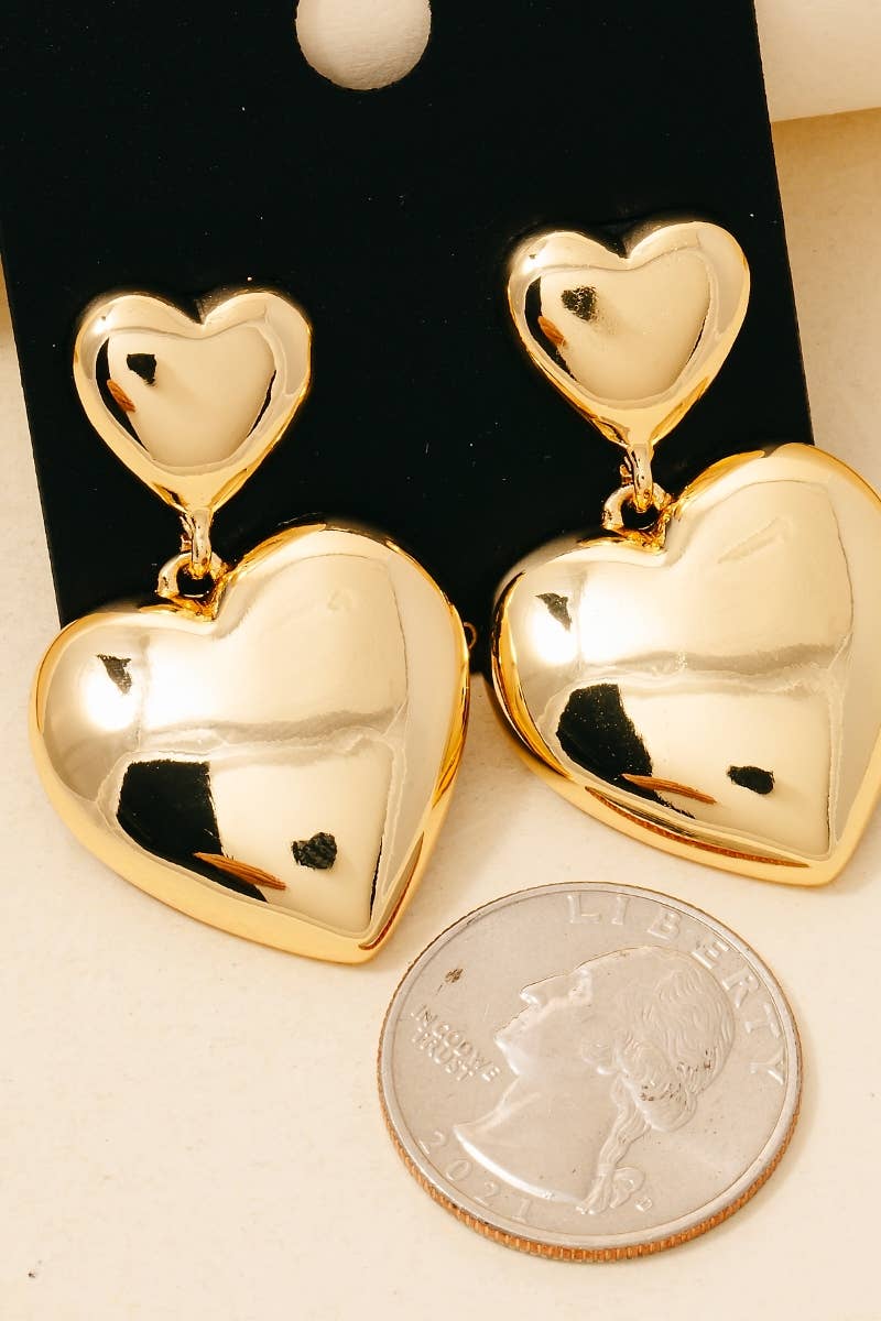 A pair of shiny gold heart-shaped drop earrings with a smaller heart above a larger one, displayed next to a silver quarter coin for scale on a light beige surface.