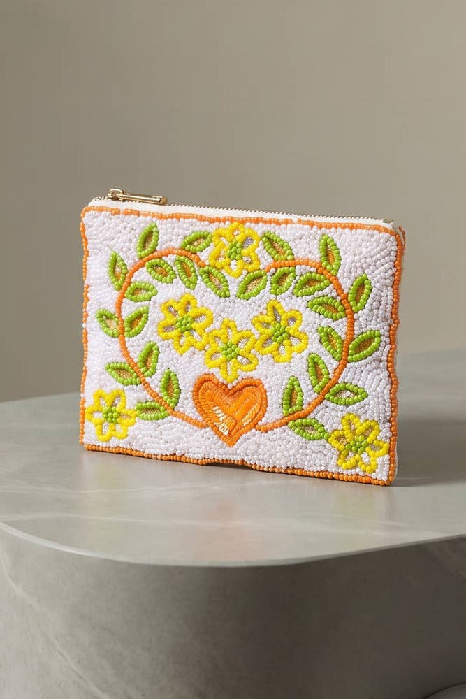A small quilted zip pouch with a white background, featuring a yellow floral heart design with green leaves and an orange heart in the center, displayed on a gray marble table against a neutral gray wall.