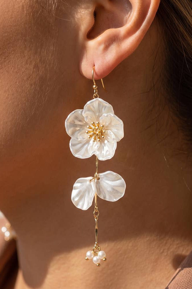 A close-up of a woman's ear wearing a dangling flower earring. The earring features a large white flower with a gold center, a smaller white flower below, and a dangling gold chain with tiny pearl-like beads at the end.