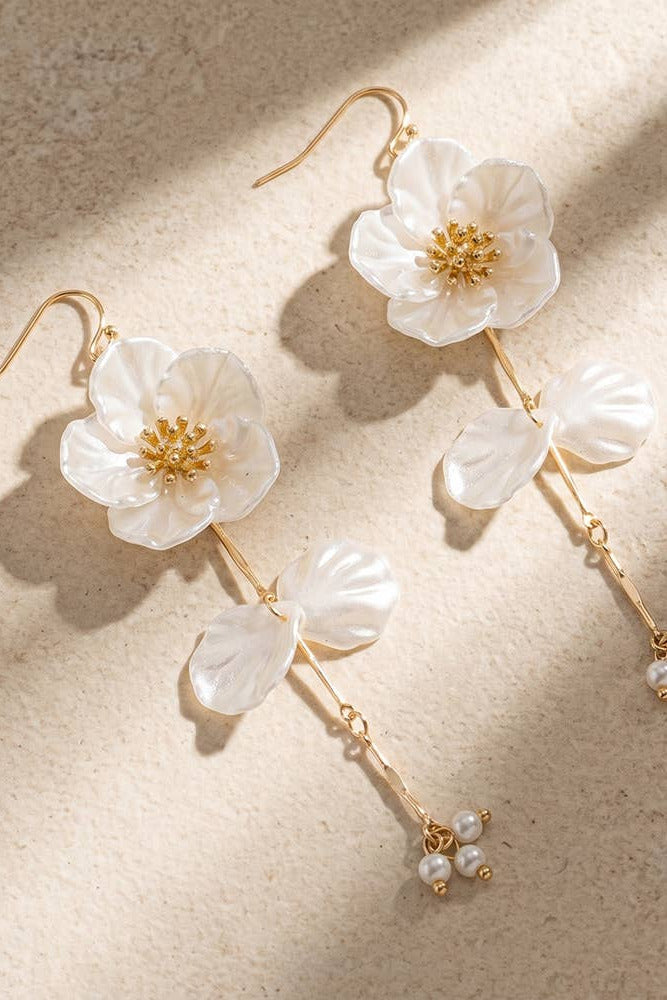 A pair of dangling earrings with large white flower charms, gold hooks and stems, and small pearl-like bead drops, photographed on a warm beige textured surface with diagonal shadows.