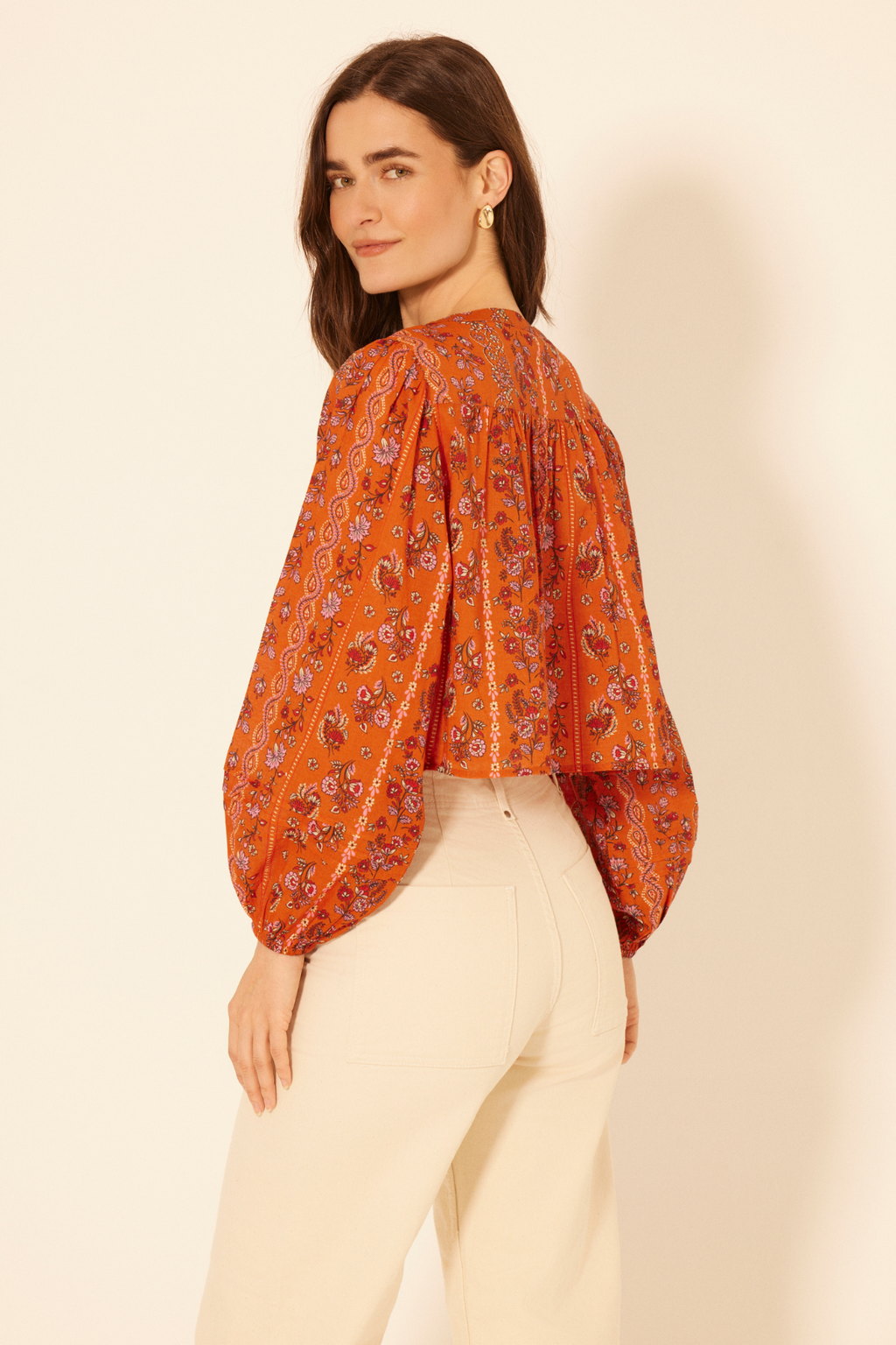 Woman wearing an orange floral blouse and white pants against a beige background