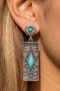 A close-up of a woman's ear wearing a silver dangle earring with a turquoise stone and engraved floral details.