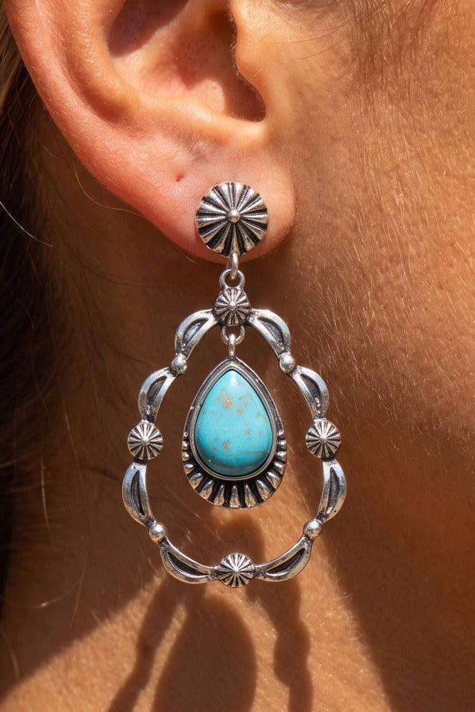 A close-up of a person's ear wearing a silver dangle earring with a turquoise teardrop stone and ornate silver settings.