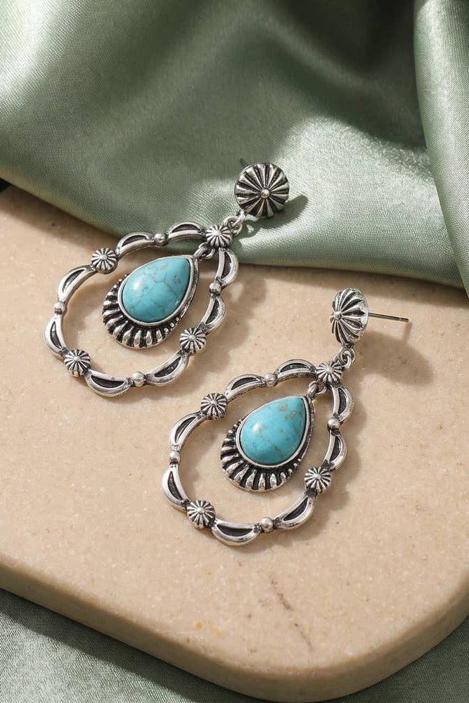 A pair of silver-toned drop earrings with turquoise teardrop-shaped stones and ornate filigree details, displayed on a beige stone slab with draped sage green satin fabric in the background.