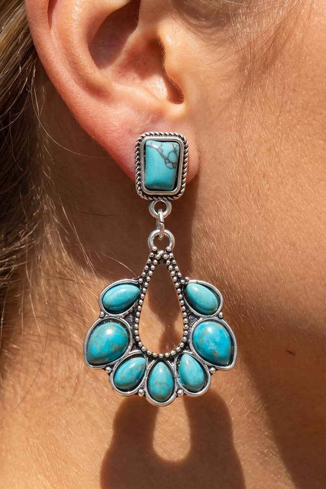 A close-up of a person's ear wearing a dangling turquoise earring with a rectangular top stone and a larger scalloped teardrop-shaped cluster of turquoise stones in silver-toned metal.