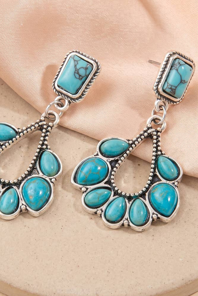 A pair of silver-toned dangle earrings with rectangular turquoise stones at the top and a fan-shaped cluster of teardrop turquoise stones below, displayed on a beige surface with peach fabric.