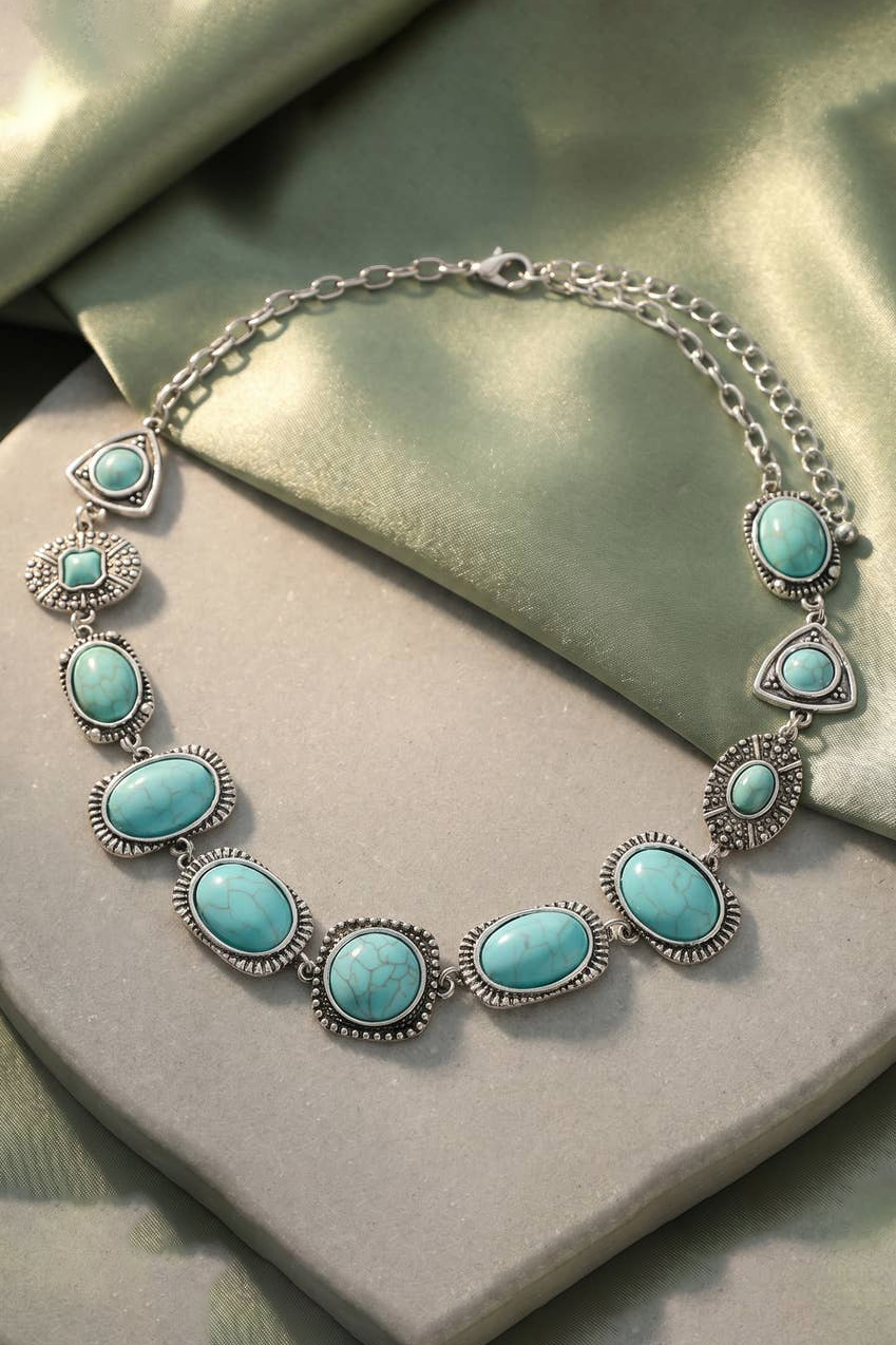 A silver-toned necklace with multiple oval and teardrop-shaped turquoise-blue stones, displayed on a light gray block with a green satin fabric background.